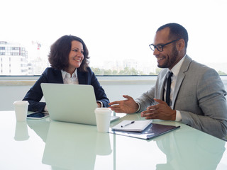 Happy coworkers discussing new business software. Business man and woman sitting at meeting table with open laptop, talking and smiling. Business software concept