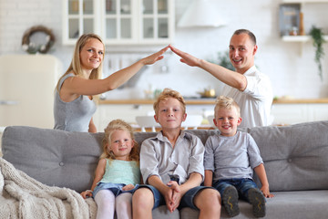 Happy full family with three kids sitting on sofa, mom and dad making roof figure with hands arms over heads. New building residential house purchase apartment and housing concept.