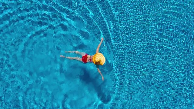 View from the top as a woman in a red swimsuit and a big yellow hat swims in the pool