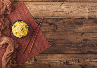 Black bowl with boiled organic basmati vegetable rice with wooden chopsticks on brown placemat with linen towel. Yellow corn and green peas with paprika slices. Space for text