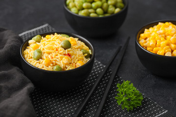 Black bowl with boiled organic basmati vegetable rice with black chopsticks on placemat with linen towel. Yellow corn and green peas with paprika slices.