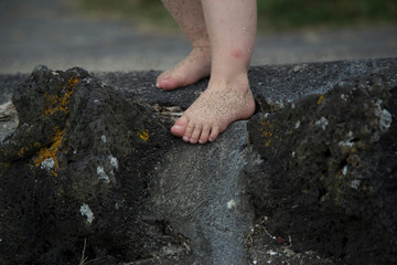 Toddler climbing down rock wall at beach with sandy feet. Could be boy or girl, gender neutral. Rocks have moss and lichen, rustic. Pt Chevalier beach, Auckland. Innocent fun, kiwi kid, nostalgic.