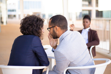 Employees gossiping about young female colleague. Business man and woman whispering, African American employee sitting in background. Office rumors concept