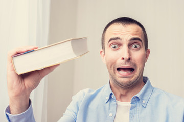 Student in shock because of a very thick book, guy does not like to read and learn, man looks at the camera in horror. The concept of heavy loads in education, toned