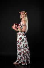 full length portrait of a blonde girl wearing a long white and floral dress, wearing a flower crown. Standing pose  on a black studio background.
