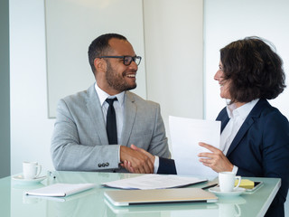Happy businesspeople closing deal. Business man and woman sitting at meeting table, holding documents and shaking hands. Contract or handshake concept
