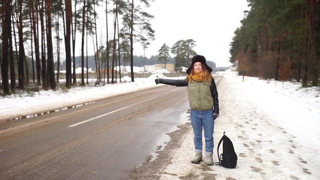 Beautiful red-haired girl hitchhiking on the road. Girl winter is worth one on the road. The girl in the hat with earflaps hitchhiking in the woods.