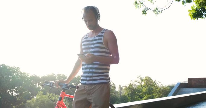 Young handsome male with colorful bicycle and headphones listen to music and ride by the summer streets, 4k futage 
