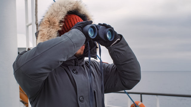 Adventurer in Warm Jacket Standing on Ship and Looking through Binoculars. It is Windy
