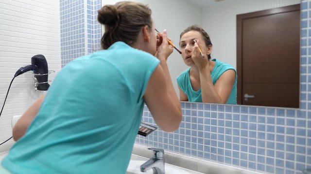 Pretty woman does eyebrow makeup with eye shadow and makeup brush in front of bathroom mirror