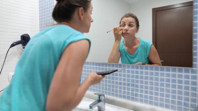 Pretty woman doing eye makeup with eyeshadow and makeup brush in front of bathroom mirror