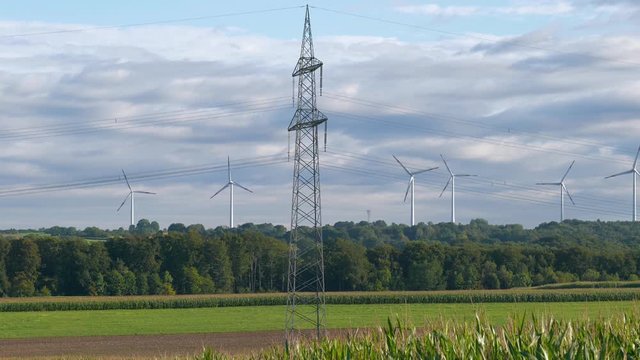 Strommast mit im Hintergrund stehenden und sich drehenden Windr&auml;dern in l&auml;ndlicher Umgebuung mit Feldern und Wald bei leicht bew&ouml;lktem Himmel