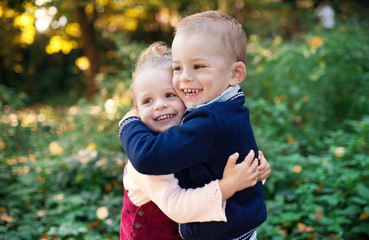 Twin toddler sibling boy and girl standing in autumn forest, hugging.