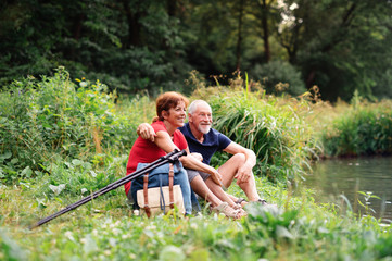 Naklejka na ściany i meble Senior tourist couple with backpacks on a walk in nature, sitting by lake.