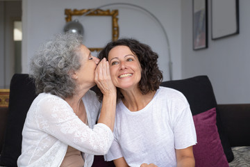Mother whispering something to daughter. Happy senior mother sharing secret with smiling adult daughter at home. Closeness concept