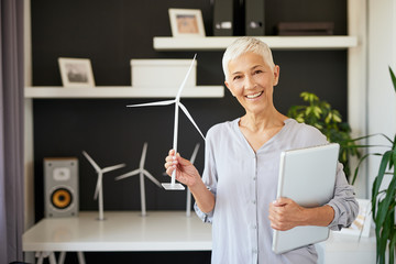Attractive smiling Caucasian senior woman dressed casual and with short gray hair standing in office, holding laptop and windmill. Sustainable concept.