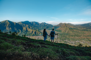 Obraz premium Young couple with backpacks hiking in the mountains and enjoying valley view. Back view of young tourist couple with backpacks, athletic man and slim girl stand holding hands on rocky mountai