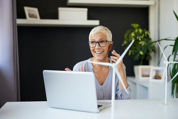 Gorgeous Caucasian senior woman talking on the phone while sitting in office. On desk are are windmill models and laptop.Start up business concept.