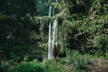 Rear view of young woman standing in front of waterfall. Female tourist with her arms outstretched looking at waterfall.
