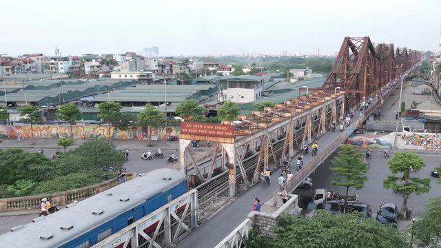 A passenger train carriage emerges from the structure of the Long Bien Bridge in Hanoi Vietnam. It is filmed from a high angle showing the sprawling city and busy traffic of Hanoi, capital of Vietnam