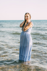 Caucasian beautiful pregnant woman in casual dress stands knee-deep in the sea. The clothes are wet, and the woman smiles. The period of expectation of a child.