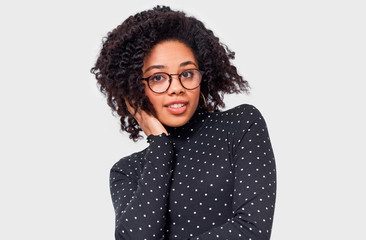 Pretty dreamy dark-skinned young woman dressed in black with white dots blouse, holding hands on head, feel happy. African American female smiling broadly, wearing round eyewear posing over white wall