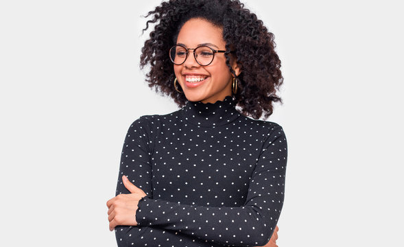 Pleased young woman dressed in black with white dots blouse, holding crossed hands, feel happy. African American female smiling broadly, wearing round transparent eyewear posing over white wall