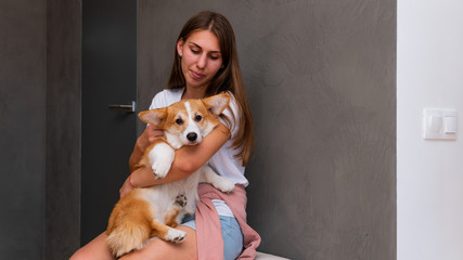 The young woman is sitting in the flat with little dog on her hand. Cute corgi Pembroke puppy on its owners hands