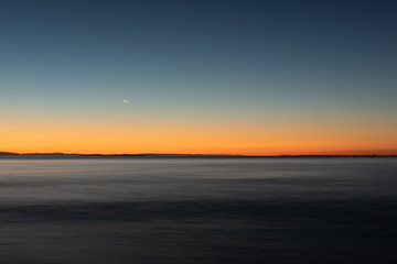 Fototapeta na wymiar A long exposure captures the crescent moon overlooking over the beautiful gradient created by the sun after it has set into the horizon on North Stradbroke Island, Queensland, Australia