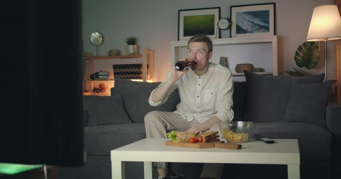 Shocked young man is watching TV breaking news drinking beer then spilling drink sitting on couch at night at home. Human reaction and television concept.