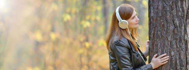 Banner. A pretty red-haired girl in white headphones in a black leather jacket looks at a tree in...