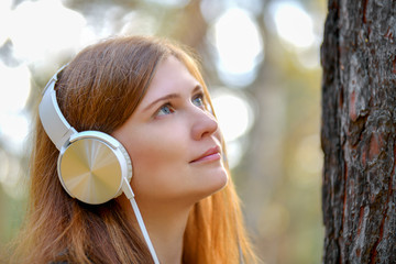 Portrait. Cute red-haired girl in white headphones looking at a tree in the woods. Girl with...