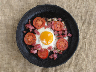 Fried eggs with sausage and tomatoes in a cast-iron frying pan against the background of burlap.  View from above.
