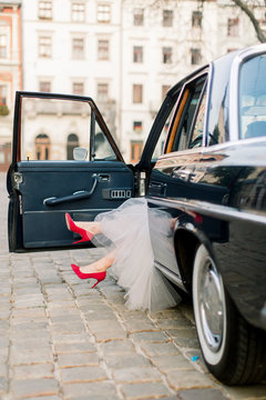 Legs of pretty bride woman in red high-heals sitting in black retro car, old buildings of the city on the background.