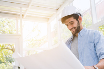 Engineer man(caucasian or european) with a beard, wearing a white safety hat, wearing a blue shirt standing looking to blueprint of the hand with a happy smile in room on sunlight  background. 