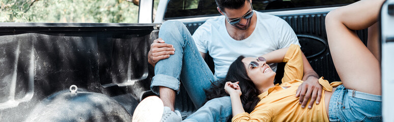 Obraz na płótnie Canvas panoramic shot of cheerful man in sunglasses sitting in car trunk near girl