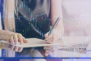 A woman hands writing information about stock market in notepad. Forex chart holograms in front. Concept of research. Double exposure