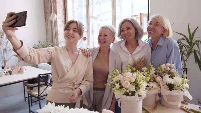 Pan shot of young Caucasian woman standing with phone in her hands and making selfie with three happy middle-aged women standing nearby in workshop