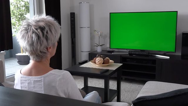 A Middle-aged Woman Sits On A Sofa In An Apartment Living Room And Watches TV With A Green Screen - View From Behind