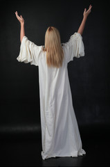 full length portrait of blonde girl wearing long white flowing robe. standing pose against a black studio background.