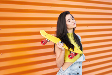 Peaceful sensual brunette girl with long hair and closed eyes wearing yellow top and blue denim overalls posing with trendy yellow skateboard with red wheels on orange wall background.