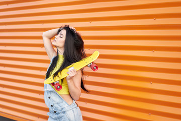 Happy joyful sensual brunette girl with long hair and closed eyes wearing yellow top and blue denim overalls posing with trendy yellow skateboard with red wheels on orange wall background