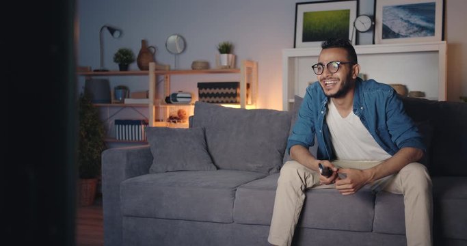 Young Arabian man is watching TV laughing having fun sitting on sofa in dark house enjoying calm evening at home. People and modern entertainment concept.