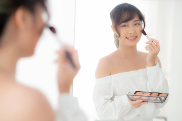 Beauty portrait young asian woman smiling with face looking mirror applying makeup with brush cheek in the bedroom, beautiful girl holding blusher, skin care and cosmetic table fashion concept.
