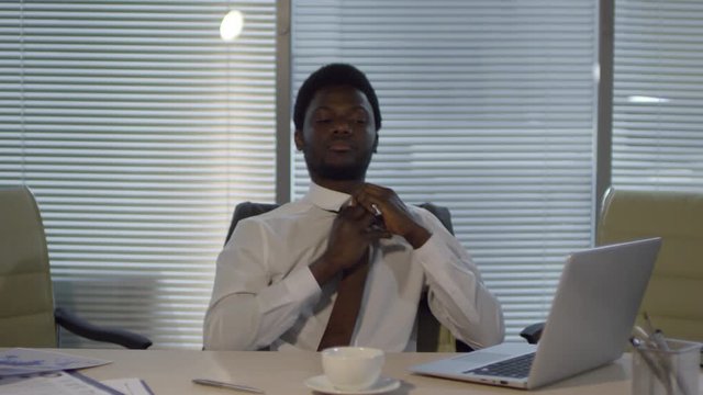 Chest-up shot of Afro-American corporate associate sitting alone in office at night and working on laptop, then rubbing face, loosening tie, unbuttoning white shirt and continuing to type
