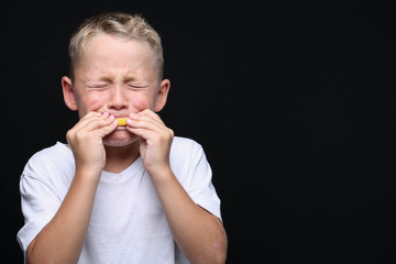 Little, blond boy is eating a piece of a lemon in front of black background and making a facial...