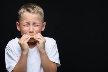 Little, blond boy is eating a piece of a lemon in front of black background and making a facial...