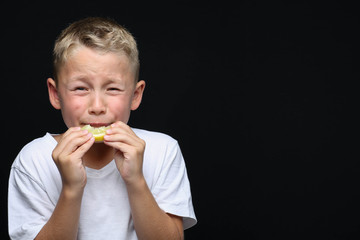 Little, blond boy is eating a piece of a lemon in front of black background and making a facial...