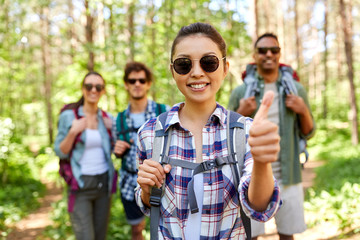 travel, tourism, hike and people concept - group of friends with backpacks and asian woman showing thumbs up gesture in forest