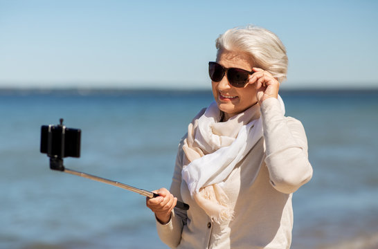old people and leisure concept - happy smiling senior woman taking picture by smartphone and selfie stick on beach in estonia